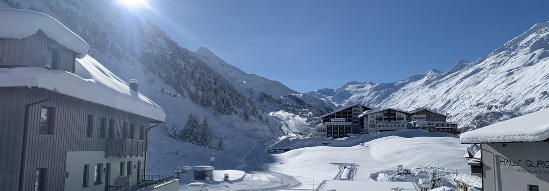 A snow-covered mountain landscape with a bright blue sky and the sun shining over the peaks. In the foreground, modern buildings can be seen. - Haus Verwall Appartements - Obergurgl 