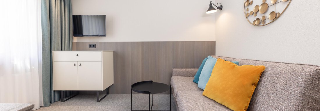 A modern room with a gray sofa and colorful cushions. Next to the sofa, there is a small table and there is a window with curtains. - Haus Verwall Appartements - Obergurgl 