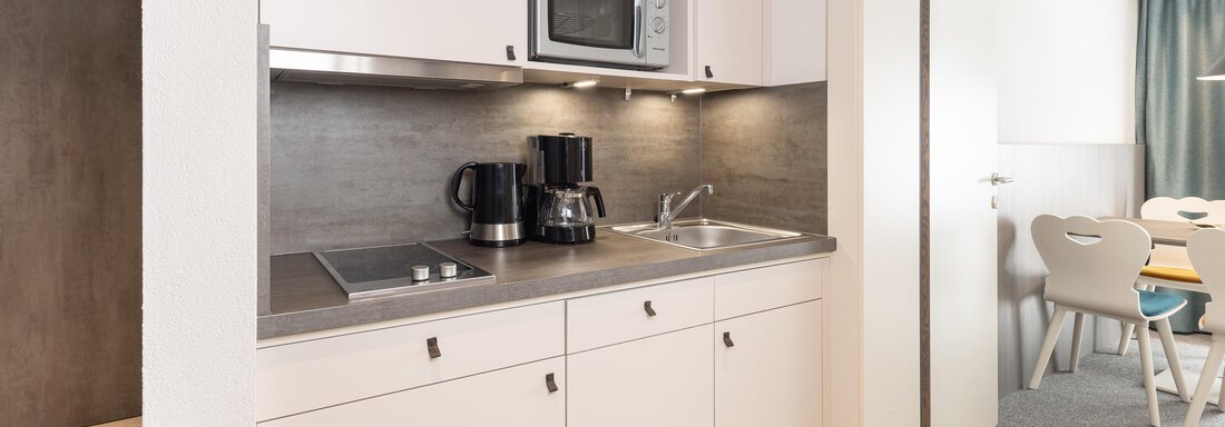 A modern kitchenette with light cabinets, stove, and microwave. In the background, a bright living area with several chairs can be seen. - Haus Verwall Appartements - Obergurgl 