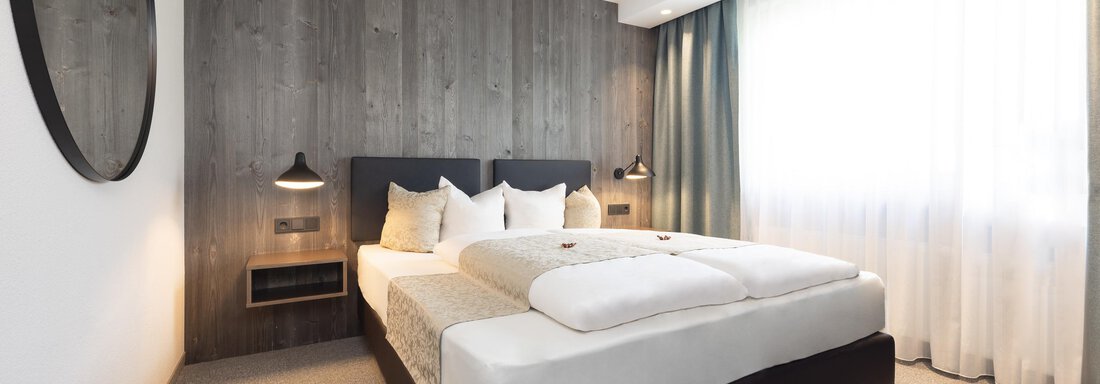 A modern bedroom with a double bed and wooden panels on the wall. Large windows provide plenty of daylight. - Haus Verwall Appartements - Obergurgl 