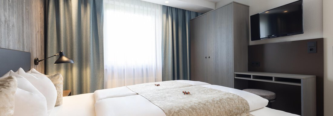 A modern hotel room with a large double bed and soft colors. The windows are draped with curtains and a television is mounted on the wall. - Haus Verwall Appartements - Obergurgl 