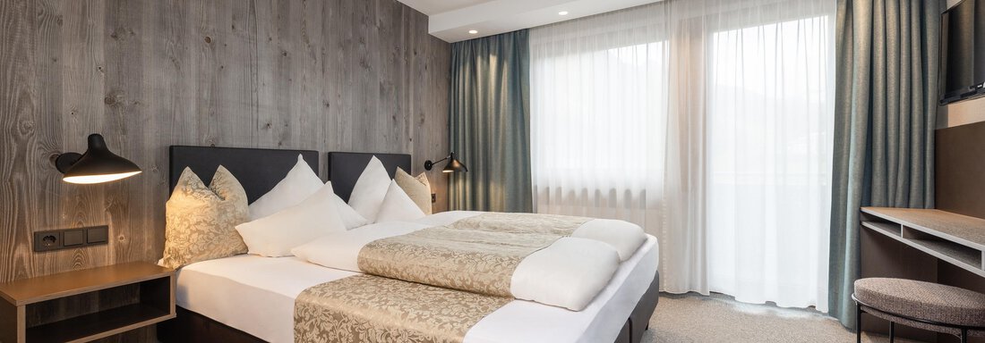 A modern hotel room with a large double bed and wooden paneling.  
Plenty of light pours in through the windows, making the room feel cozy and inviting. - Haus Verwall Appartements - Obergurgl 