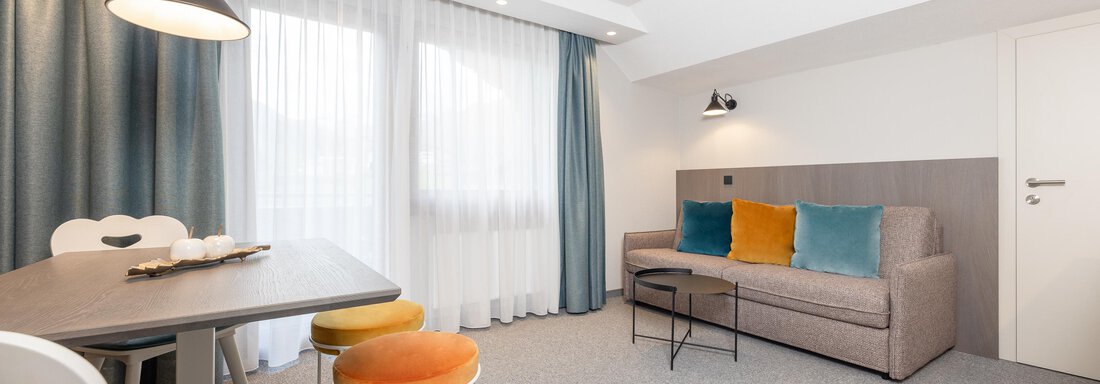 A modern living room with a comfortable sofa and a dining table. The large windows let in plenty of daylight, and the colorful cushions give the space a cheerful accent. - Haus Verwall Appartements - Obergurgl 