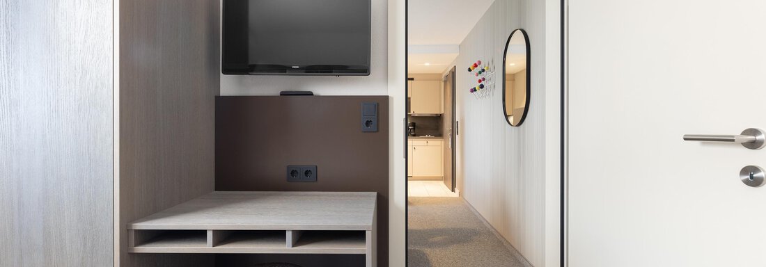 A simple room with a desk, a chair, and a television on the wall. In the background, there is a hallway with another door. - Haus Verwall Appartements - Obergurgl 