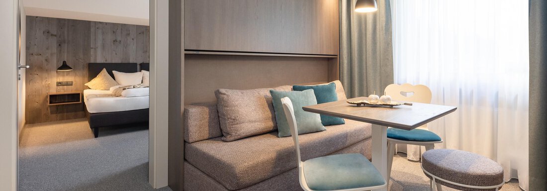 A modern living and dining room with a cozy sofa and a table. The space is bright and cheerful with large windows and simple furniture. - Haus Verwall Appartements - Obergurgl 