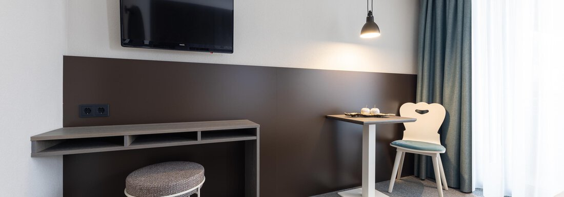 A modern room with a television, a desk, and a cozy seating area. The room has large windows with curtains and is designed to be bright and inviting. - Haus Verwall Appartements - Obergurgl 