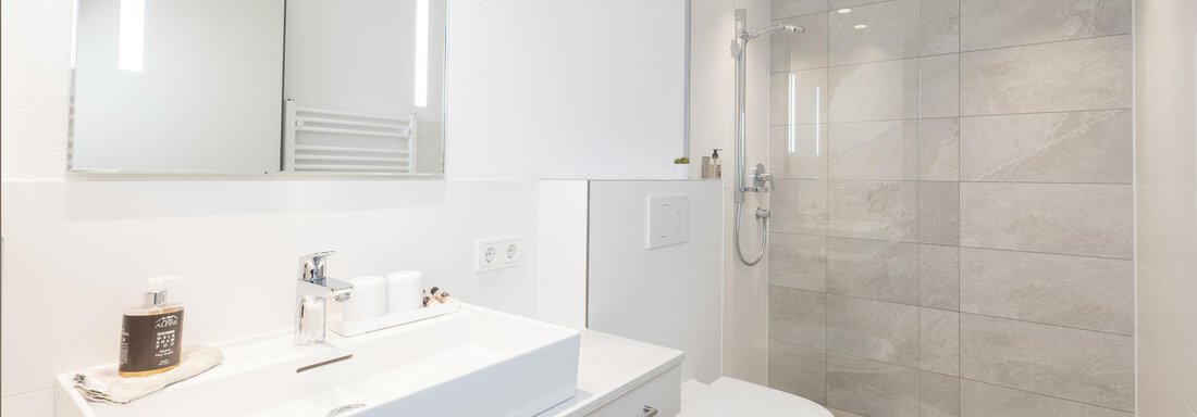 A modern bathroom with a white sink and a large mirror. The shower is transparent and the walls are decorated with light tiles. - Haus Verwall Appartements - Obergurgl 