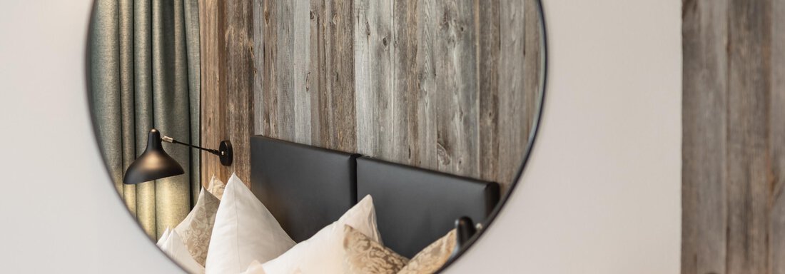 A round mirror reflects a stylish bedroom with a wooden wall design. Bright pillows are on the bed, and there are two modern lamps. - Haus Verwall Appartements - Obergurgl 