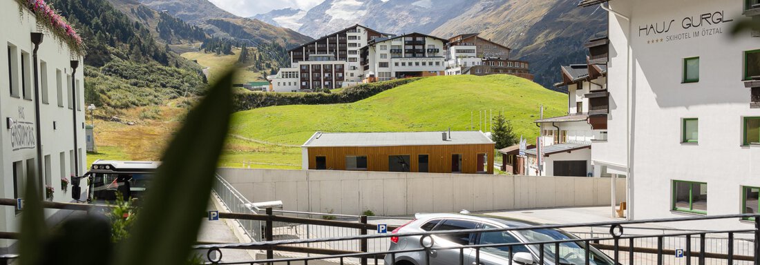 A tranquil mountainous landscape with modern buildings and a clear sky. In the foreground, some cars and plants can be seen. - Haus Verwall Appartements - Obergurgl 