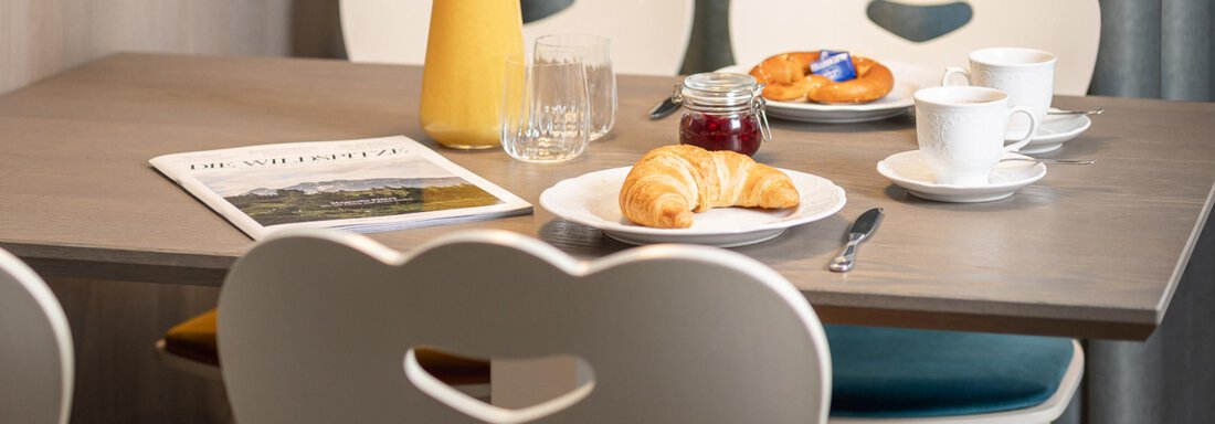 A cozy breakfast table with croissants, a cup of coffee, and freshly squeezed orange juice. In the background, there are chairs with a heart-shaped design. - Haus Verwall Appartements - Obergurgl 