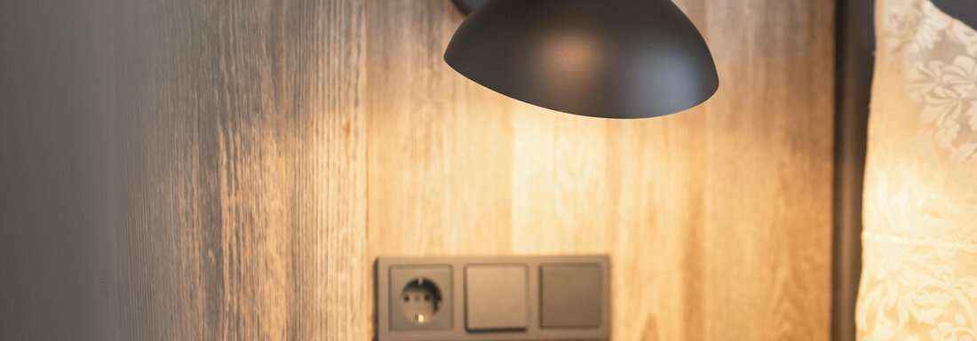 A modern lamp on a wood wall above a nightstand. The switches and outlet are clearly visible and harmonize with the minimalist design. - Haus Verwall Appartements - Obergurgl 