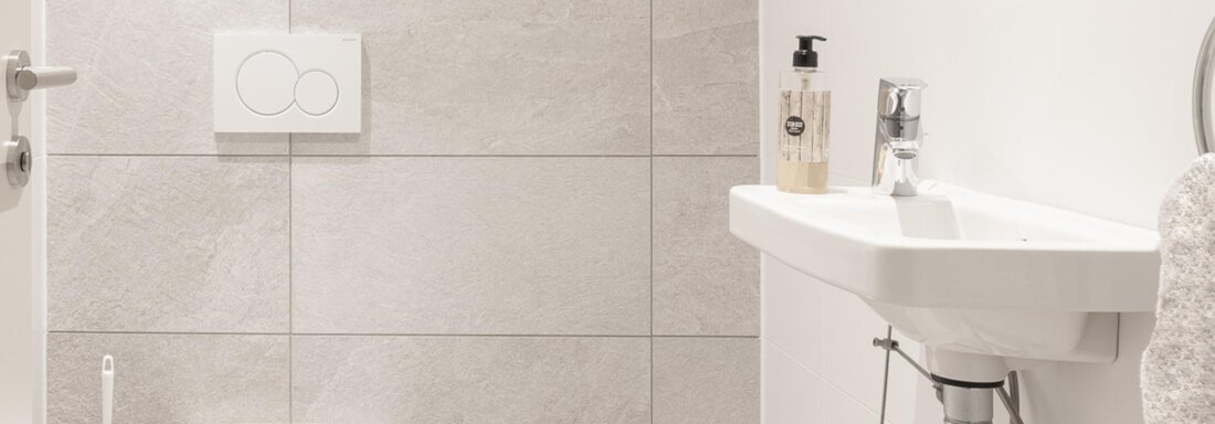 A modern bathroom with a white toilet and a small sink. The walls are light-tiled, and there are some toiletries on the shelf. - Haus Verwall Appartements - Obergurgl 