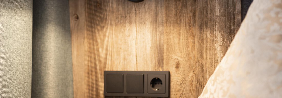 A modern bedside lamp hangs over a wooden panel. Below it is a socket with two switches. - Haus Verwall Appartements - Obergurgl 
