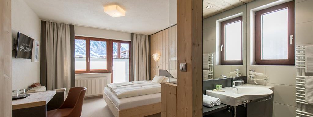 Komfortzimmer Neu ! A modern hotel room with a double bed and a workspace. The room features large windows with a view outside and an adjoining bathroom. - Hotel-Garni Pirchhütt - Obergurgl