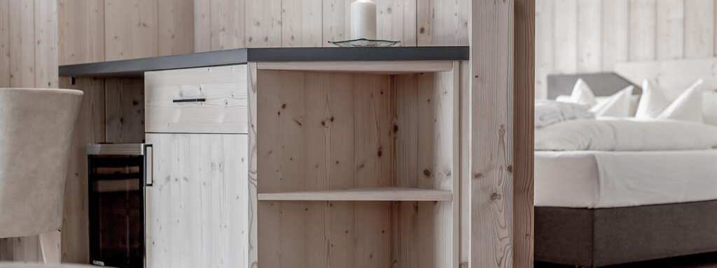 A modern kitchen corner made of light wood with a candle on the table. In the background, a comfortable bed can be seen in a stylish room. - Hotel-Garni Pirchhütt - Obergurgl 
