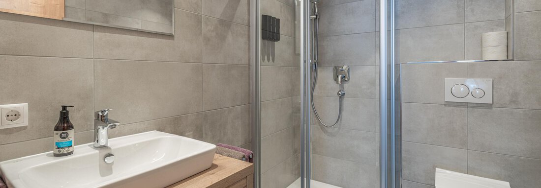 A modern bathroom with a shower and an elegant wooden sink unit. The walls are clad with gray tiles, and there is a minimalist toilet. - Mein Gaisberg - Obergurgl 