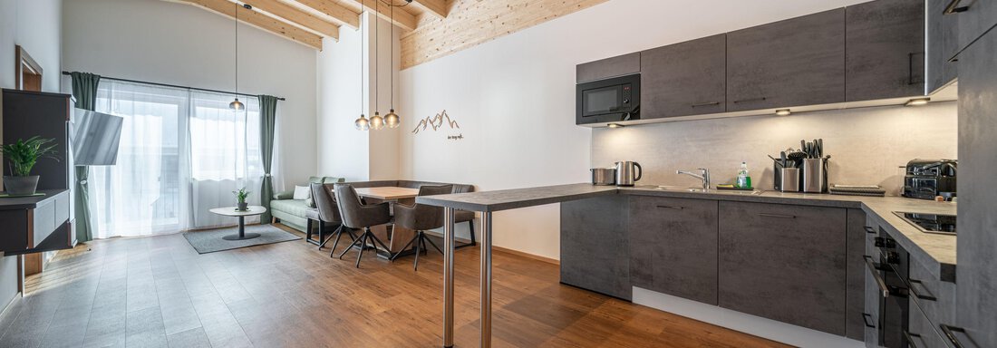 A modern kitchen with wooden ceilings and an open living area. The space is bright and friendly with a large window and a dining table. - Mein Gaisberg - Obergurgl 