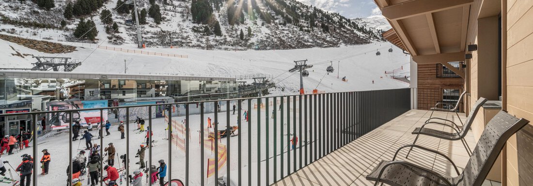 A terrace with two chairs and a view of a busy ski resort. In the background, snow-covered mountains and a sunny atmosphere can be seen. - Mein Gaisberg - Obergurgl 