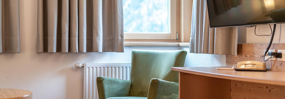 A cozy room with a green armchair and a table. A large television hangs on the wall next to a window with curtains. - Schönblick Gurgl - Obergurgl 