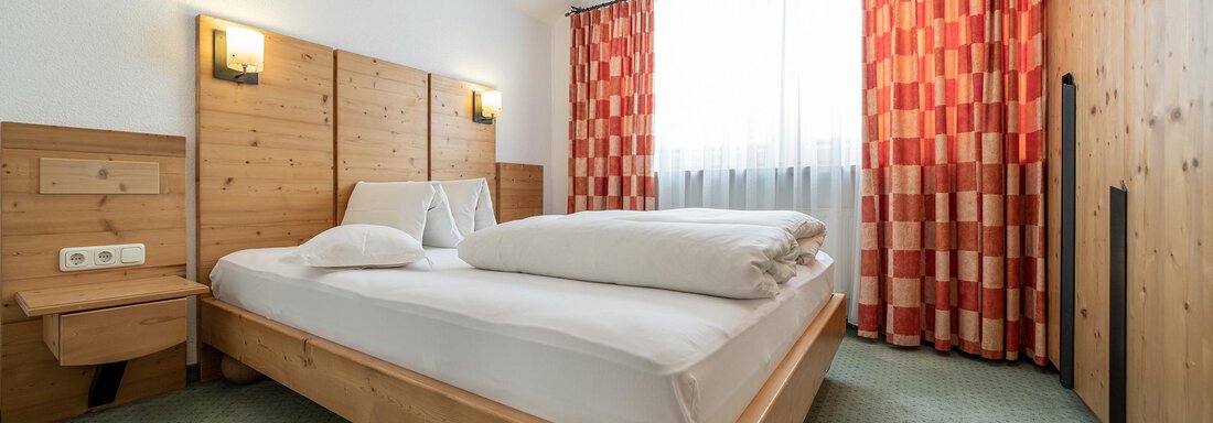 A cozy bedroom with a wooden bed and two pillows. The windows are decorated with colorful curtains and there is a green carpet. - Schönblick Gurgl - Obergurgl 