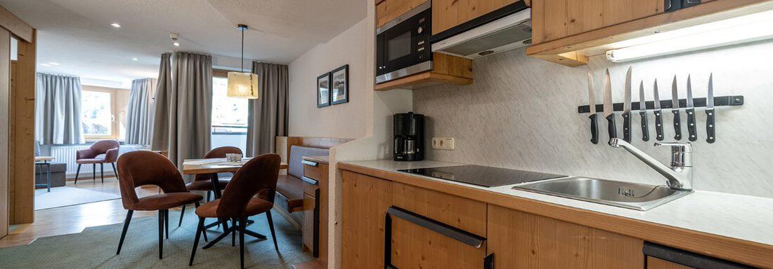 Küche Appartement "6" A cozy kitchen with wooden cabinets and a modern stove. In the background, there is a dining and living area with comfortable chairs. - Schönblick Gurgl - Obergurgl