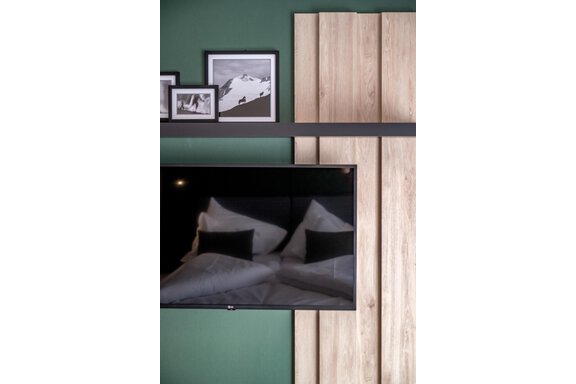 A modern living room with a television and a wooden shelf. The walls are painted in a calming green shade, and there are some framed pictures visible. - The Moss - Your unique hideaway - Obergurgl 
