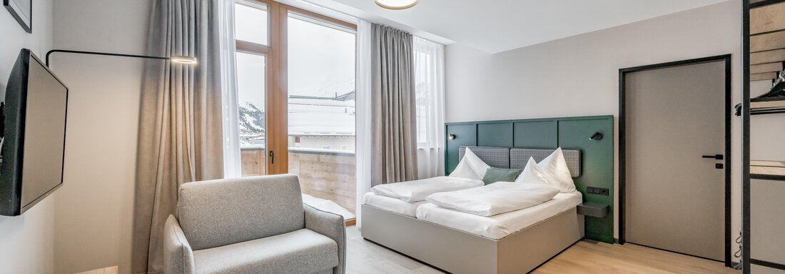A modern hotel room with a large bed and a cozy armchair. Large windows let in plenty of light and offer a view outside. - The Moss - Your unique hideaway - Obergurgl 