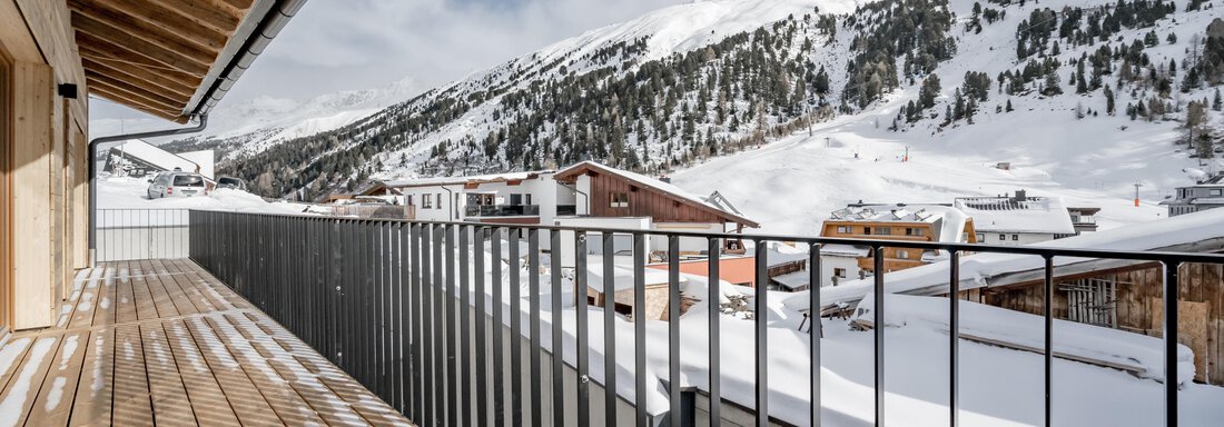 A balcony with a wooden floor and metal railing that offers a view of snow-covered mountains. In the background, houses and a clear mountain landscape can be seen. - The Moss - Your unique hideaway - Obergurgl 