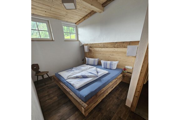 d7295c16-c70a-499d-a46e-f7f882a24b83 Ein gemütliches Schlafzimmer mit einem Holzbett und blauen Bettwäsche. Es gibt ein Fenster, das Tageslicht hereinlässt, und rustikale Möbel. - Almhof Mareil - Ochsengarten