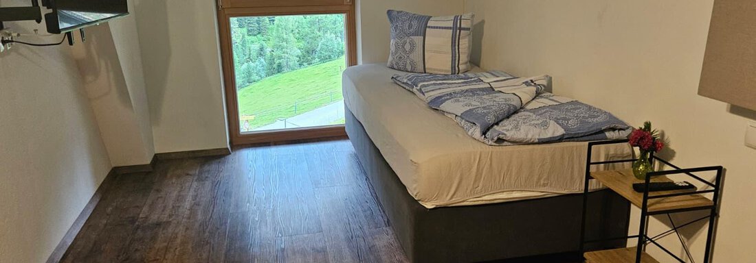 Ein gemütliches Schlafzimmer mit einem Einzelbett und einem Fernseher. Große Fenster bieten einen Blick auf die grüne Landschaft. - Almhof Mareil - Ochsengarten