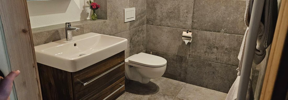 Ein modernes Badezimmer mit einer Waschgelegenheit und einem WC. Die Wände sind mit grauen Fliesen und schönen Akzenten dekoriert. - Almhof Mareil - Ochsengarten