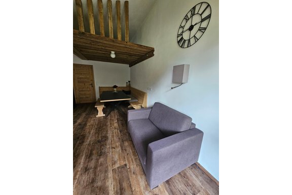 0552ce2b-d3e9-4ad7-8f1a-22f8b03ba98b Ein stilvolles Wohnzimmer mit einem grauen Sofa und einem rustikalen Holztisch. Hoch oben ist ein offenes Obergeschoss sichtbar, und an der Wand hängt eine große Uhr. - Almhof Mareil - Ochsengarten