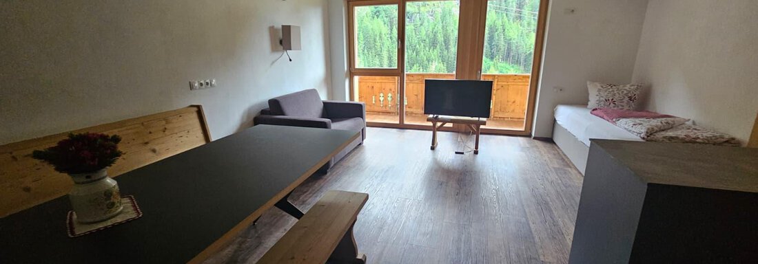 Ein modernes, helles Wohnzimmer mit großen Fenstern und Blick auf die Natur. Der Raum enthält einen Esstisch, eine Couch und einen Fernseher. - Almhof Mareil - Ochsengarten