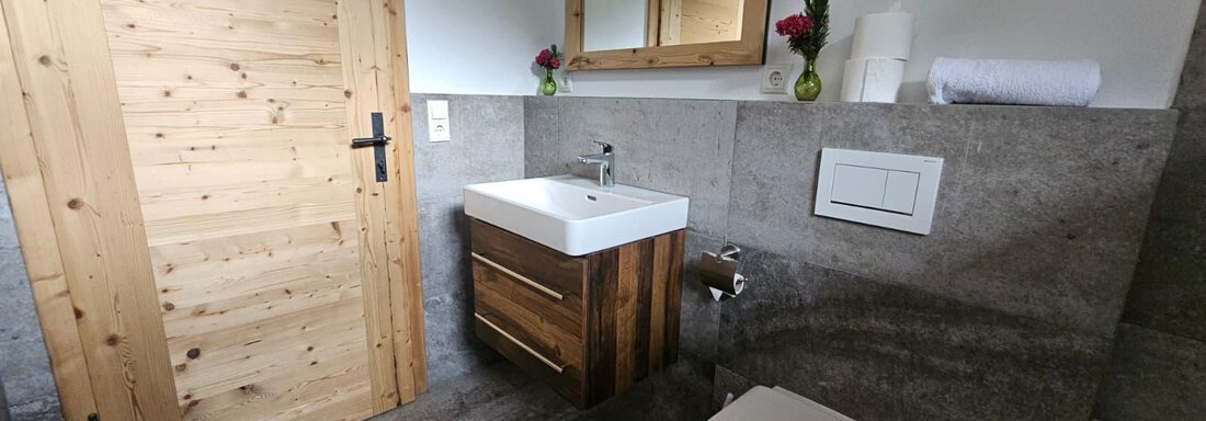 Ein modernes Badezimmer mit einem Holzwaschtisch und einem eleganten Spiegel. Die Wände sind aus grauem Stein und die Tür ist aus Holz. - Almhof Mareil - Ochsengarten