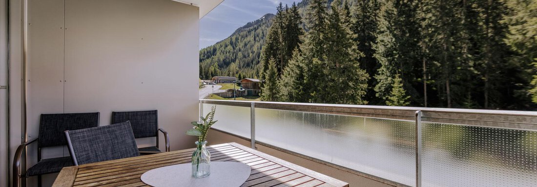 Ein Balkon mit einem Holztisch und Stühlen, umgeben von einer schönen Berglandschaft. Die Aussicht zeigt hohe Bäume und einen klaren Himmel. - Apartment Waldesruh - Ochsengarten