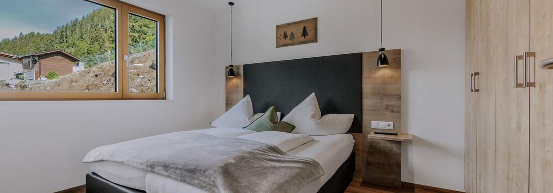 Ein modernes Schlafzimmer mit einem großen Bett und einer Holzverkleidung. Durch das Fenster sieht man eine schöne Waldlandschaft. - Apartment Waldesruh - Ochsengarten