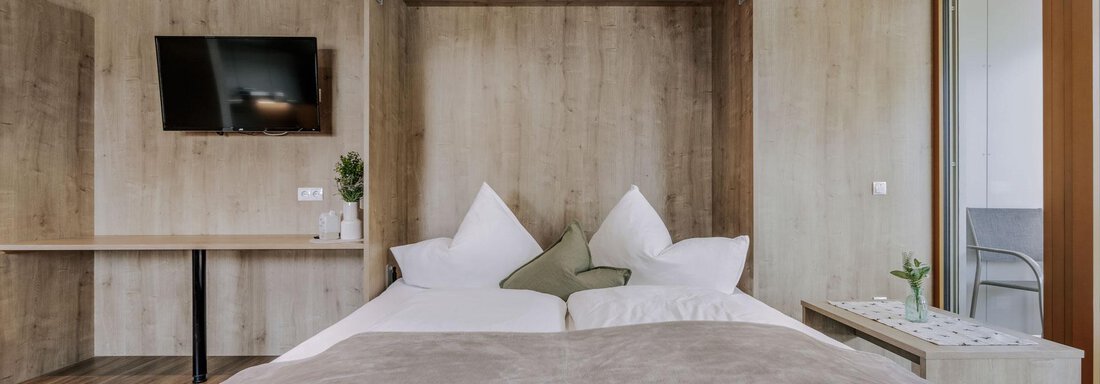 Ein modernes Schlafzimmer mit einem großen Bett und zwei Kissen. An der Wand hängt ein Fernseher, und es gibt einen kleinen Tisch sowie eine Terrasse mit einem Stuhl. - Apartment Waldesruh - Ochsengarten