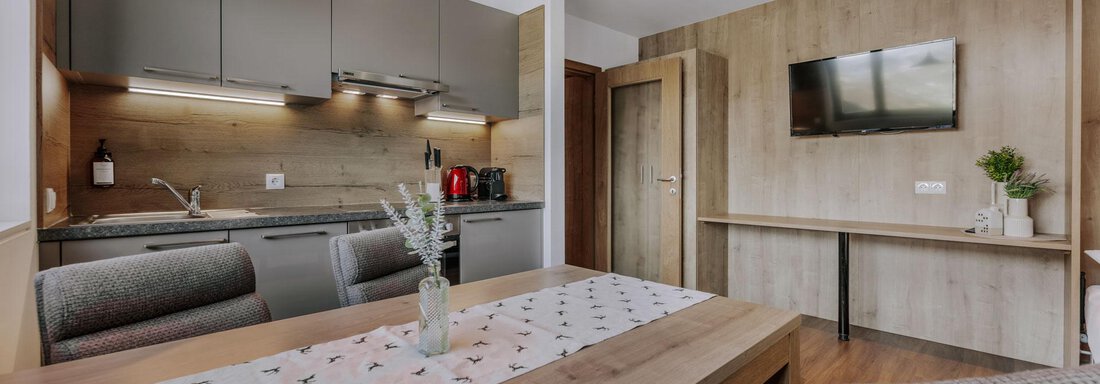 Eine moderne Küche mit einem Esstisch und bequemen Stühlen. An der Wand hängt ein Fernseher, und die Einrichtung ist in warmen Holztönen gehalten. - Apartment Waldesruh - Ochsengarten