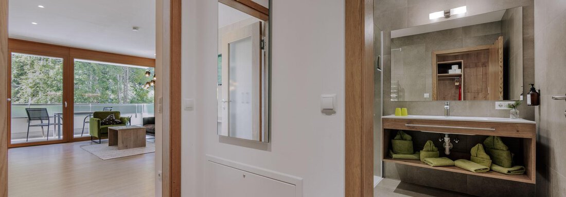 Ein moderner Flur mit Blick auf ein Badezimmer. Der Raum ist hell und freundlich, mit Holz- und Grautönen sowie einem großen Spiegel. - Apartment Waldesruh - Ochsengarten