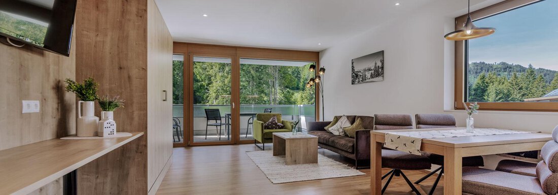 Ein modernes Wohnzimmer mit einem großen Fenster, das einen Blick auf die Natur bietet. Es gibt eine gemütliche Couch, einen Esstisch und einen Fernseher an der Wand. - Apartment Waldesruh - Ochsengarten