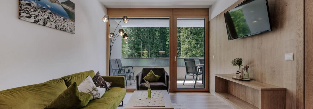 Ein modernes Wohnzimmer mit einem grünen Sofa, einem Couchtisch und einem großen Fernseher. Durch die Fenster ist eine Terrasse mit Stühlen und viel Grün sichtbar. - Apartment Waldesruh - Ochsengarten