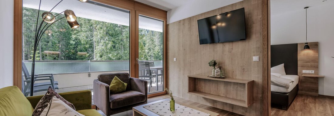 Ein modernes Wohnzimmer mit einem gemütlichen Sofa und einem Fernseher an der Wand. Große Fenster bieten einen Blick auf die Natur draußen. - Apartment Waldesruh - Ochsengarten