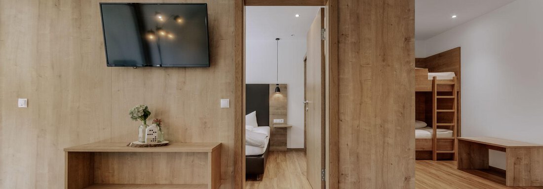 Ein modernes Zimmer mit Holzboden und einem Fernseher an der Wand. Auf der rechten Seite ist ein Bereich mit einem Etagenbett sichtbar. - Apartment Waldesruh - Ochsengarten