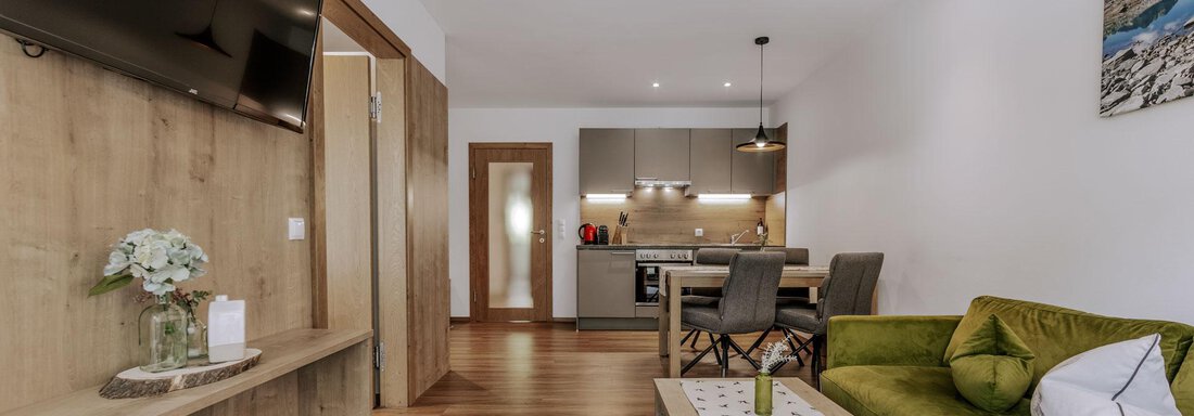 Eine moderne Wohnküche mit einem grünen Sofa und Holzboden. Der Raum ist hell und freundlich gestaltet, mit einem Fernseher an der Wand und einem Esstisch. - Apartment Waldesruh - Ochsengarten
