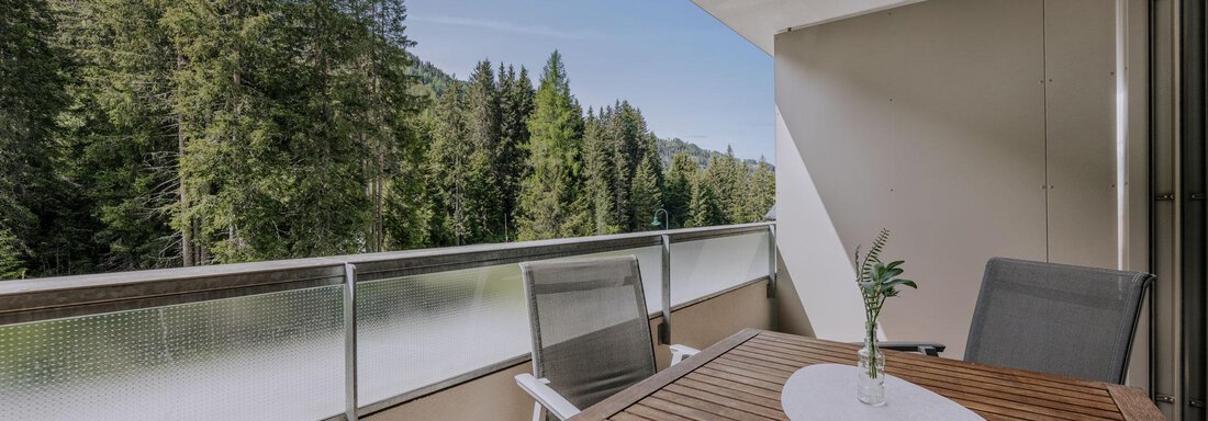 Ein moderner Balkon mit einem Holztisch und zwei Stühlen. Im Hintergrund sind hohe, grüne Bäume sichtbar. - Apartment Waldesruh - Ochsengarten