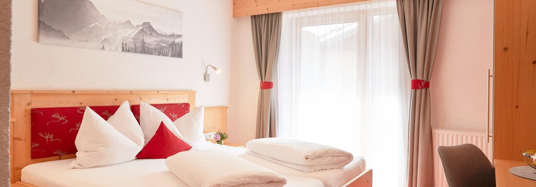 Ein gemütliches Schlafzimmer mit einem großen Bett, Holzverkleidung und einem Blick auf die Berge. Vor den Fenstern hängen helle Vorhänge, die viel Licht hereinlassen. - Gasthof Walderhof - Ochsengarten