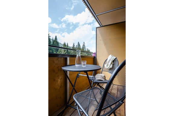 Ein kleiner Balkon mit einem runden Tisch und zwei Stühlen. Auf dem Tisch stehen eine Karaffe und ein Glas Wasser, umgeben von einer schönen Aussicht auf Bäume und blauen Himmel. - Gasthof Waldesruh - Ochsengarten