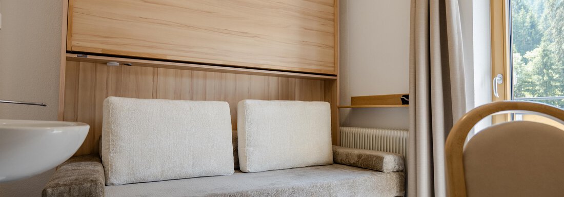 Ein gemütliches Zimmer mit einem hellen Sofa und einem Fenster, das Tageslicht hereinscheinen lässt. Die Holzverkleidung und die ruhigen Farben schaffen eine einladende Atmosphäre. - Gasthof Waldesruh - Ochsengarten