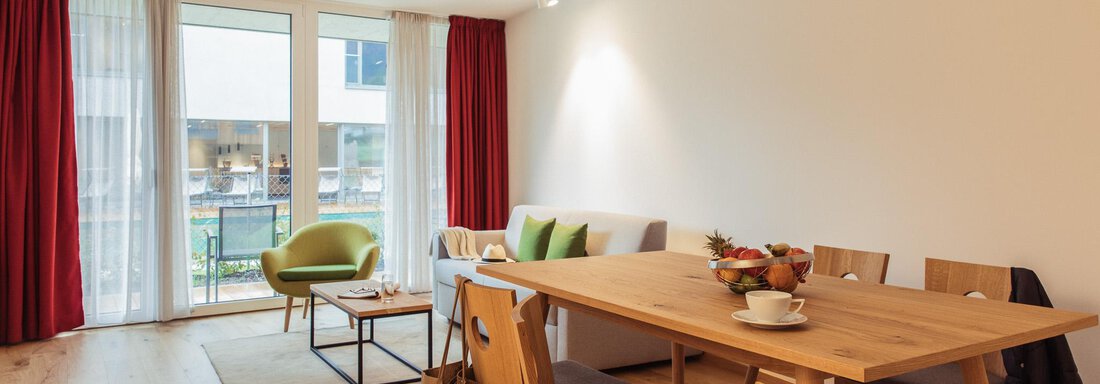 A modern living room with a dining table and chairs. Large windows let in plenty of light and offer a view outside. - All-Suite Resort Ötztal - Oetz