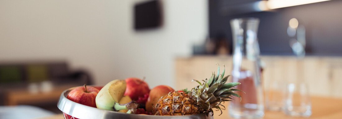 A modern kitchen with a fruit bowl on a wooden table. The bowl contains various fruits like apples, pineapples, and citrus fruits. - All-Suite Resort Ötztal - Oetz
